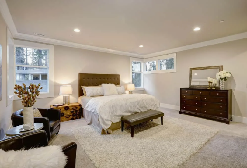 A spacious and elegantly appointed bedroom featuring neutral walls, a dark tufted headboard, a white-draped bed, and a dark wood dresser, providing wonderful inspiration for how to decorate when everything doesn't match by harmonizing contrasting tones and textures.