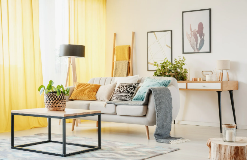 A bright and cheerful living room featuring a light gray couch with yellow and patterned throw pillows, yellow curtains, a wooden coffee table, and stylish wall art, showcasing a creative approach to how to decorate when everything doesn't match.