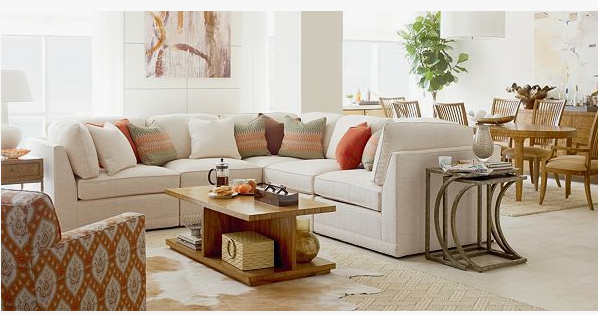 A bright and open living and dining space featuring a large beige sectional sofa adorned with patterned and solid orange throw pillows, a tiered wooden coffee table, and an adjacent dining area, creating a cohesive look on how to decorate when everything doesn't match.