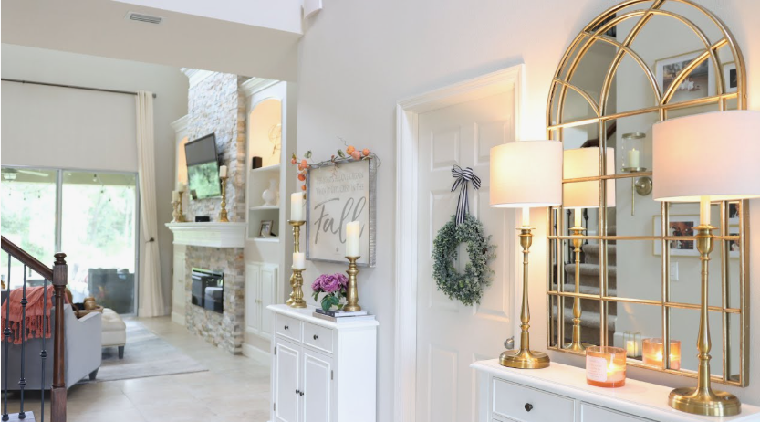 An elegantly styled home entryway featuring a large arched gold mirror, matching brass table lamps, a white cabinet with fall decor, and a view into a living room with a stone fireplace, demonstrating how to decorate when everything doesn't match.