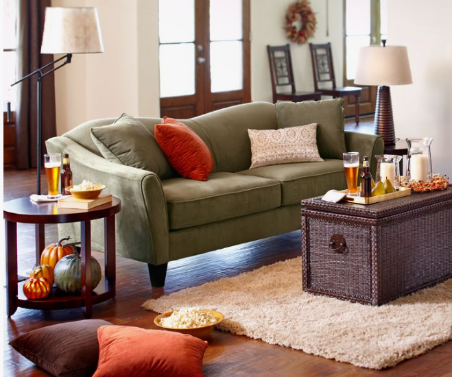 A warm, autumn-styled living room featuring an olive green sofa with orange pillows, a wicker storage chest for a coffee table, and seasonal harvest decor, showcasing how to decorate when everything doesn't match.