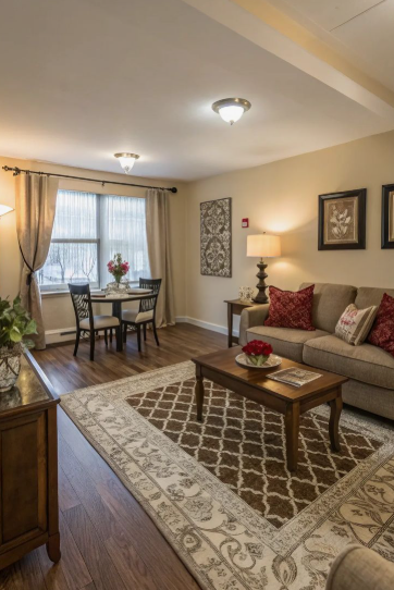 A combined living and dining room featuring a neutral sofa with red throw pillows, a dark wood coffee table, a patterned area rug, and a small dining table set by the window, demonstrating how to decorate when everything doesn't match.