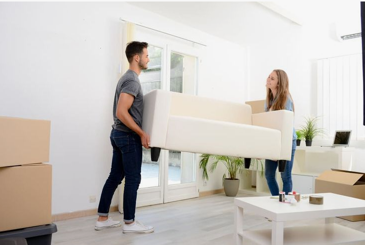 A young couple carrying a white sofa into a bright, unfurnished room with moving boxes and a coffee table, demonstrating the beginning of learning how to decorate when everything doesn't match.
