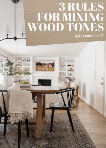 A brightly lit dining room featuring a wooden table surrounded by black chairs and a patterned rug, with a view of a living room with built-in shelves, showcasing how to decorate when everything doesn't match.