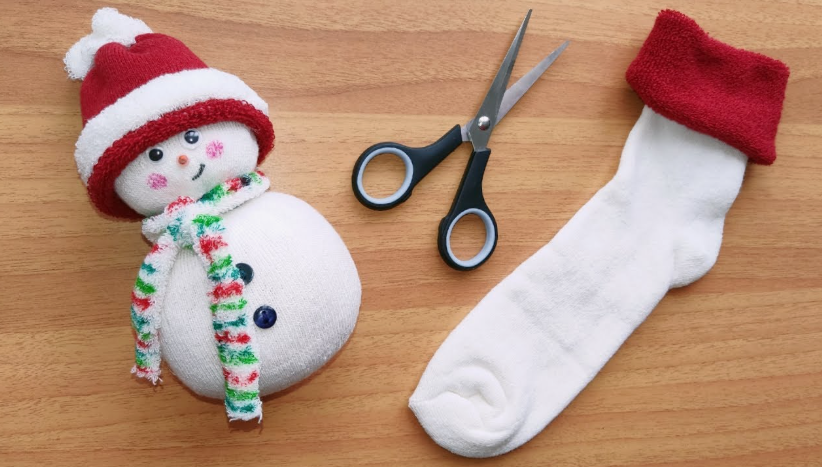 A handmade sock snowman wearing a red hat and colorful scarf, placed next to scissors and an uncrafted white sock on a wooden table, showcasing cheap DIY crafts on a budget.