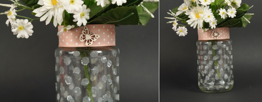 A clear glass jar painted with white polka dots, adorned with a pink ribbon and a wooden butterfly embellishment, and filled with white flowers, demonstrating cheap DIY crafts on a budget.