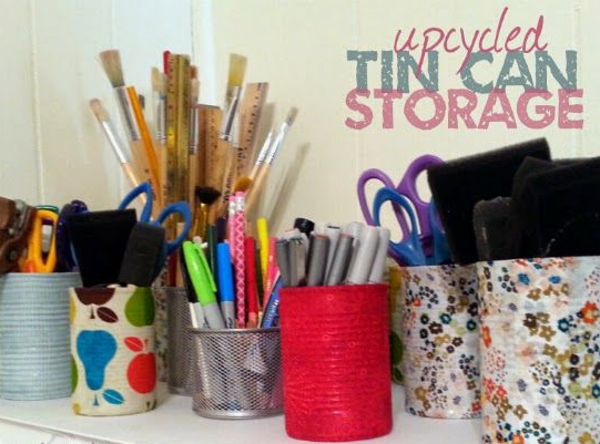 Several upcycled tin cans wrapped in patterned paper and fabric, serving as organizers for art supplies like brushes, pens, and scissors, showcasing cheap DIY crafts on a budget.