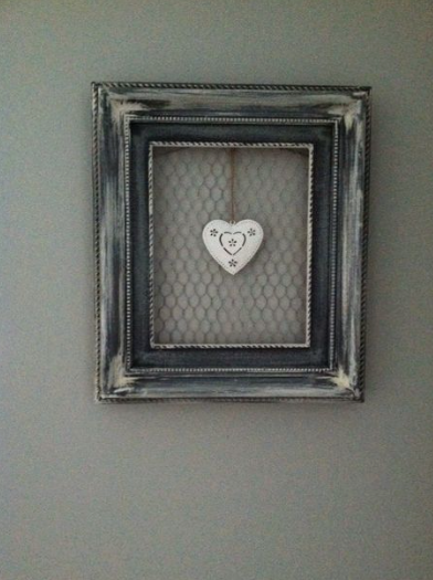 A distressed dark wooden frame fitted with chicken wire mesh and a single white decorative heart hanging from a string in the center, showcasing cheap DIY crafts on a budget.