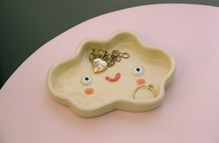 A light yellow ceramic trinket tray shaped like a smiling cloud, decorated with facial features and holding a gold locket necklace and a ring, demonstrating cheap DIY crafts on a budget.