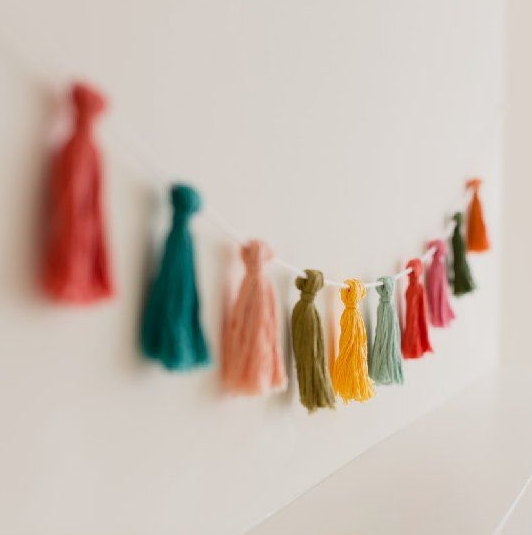 A handmade garland strung on a white cord featuring colorful yarn tassels in various bright hues hung against a plain wall, showcasing cheap DIY crafts on a budget.