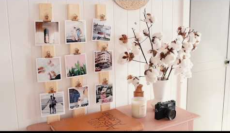 Vertical wooden slats mounted on a wall with square photos attached using clips, alongside a tabletop arrangement with a camera and cotton branch vase, showcasing cheap DIY crafts on a budget.