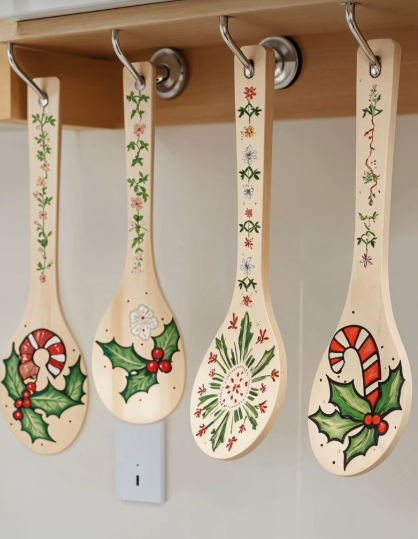 Four wooden spoons hand-painted with festive holiday designs, holly leaves, and candy canes, hanging from metal hooks under a shelf, showcasing cheap DIY crafts on a budget.