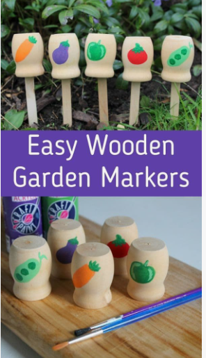 Hand-painted wooden garden markers featuring different painted vegetables on spools attached to sticks, set against a garden bed and a wooden workbench, showcasing cheap DIY crafts on a budget.