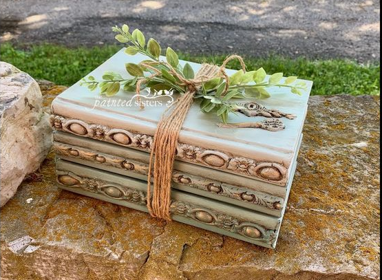 A stack of painted decorative books adorned with ornate moldings, bound with twine, and topped with green foliage and decorative keys resting on a stone surface, showcasing cheap DIY crafts on a budget.