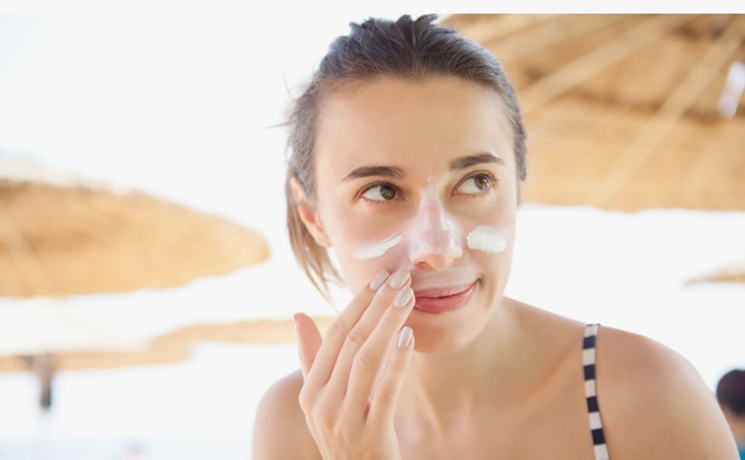 A woman applying sunscreen lotion to her face outdoors under beach umbrellas, showcasing beauty tips for women over 40.