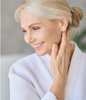 A smiling mature woman with blonde hair in a white bathrobe gently touching her neck, showcasing beauty tips for women over 40.