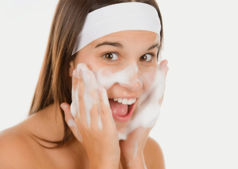 A smiling woman washing her face with rich facial cleanser foam and wearing a white headband, showcasing beauty tips for women over 40.
