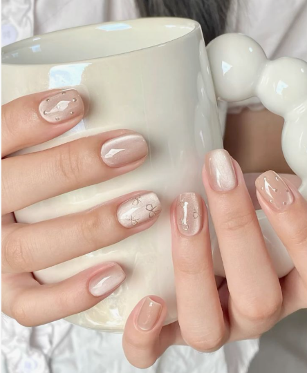 Hands holding a white ceramic mug showcasing neutral glossy office-friendly nail designs with delicate bow accents and subtle shimmer.