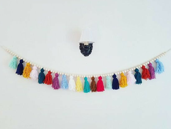 10 DIY Dorm Room Decor Ideas on a Budget That Look Amazing DIY dorm room decor ideas on a budget featuring a colorful yarn tassel and natural wood bead garland hanging on a white wall beneath a small wall sconce.