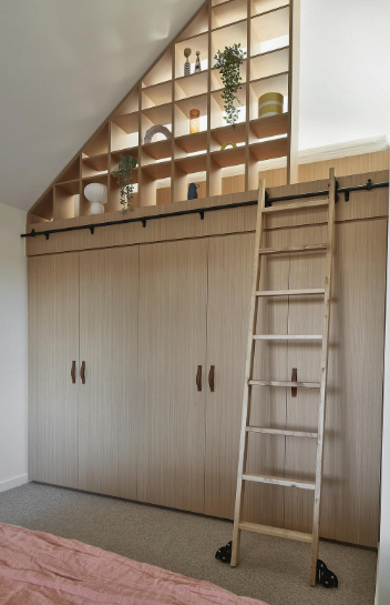 10 DIY Dorm Room Decor Ideas on a Budget That Look Amazing DIY dorm room decor ideas on a budget featuring a wall of built-in light wood wardrobes with vertical cabinet doors and leather pulls, topped by a built-in triangular cubby shelving unit with integrated lighting and decorative objects, accessible via a rolling wooden library ladder on a black track.
