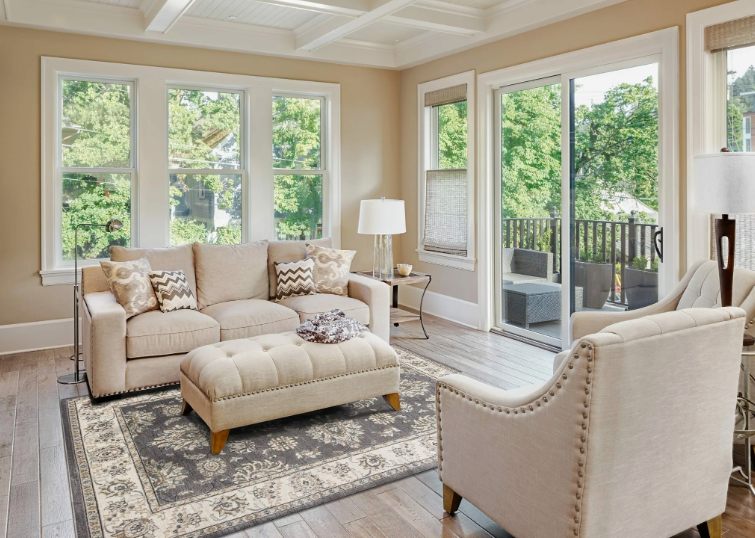 How to Make a Boring Room Look Better: 15 Simple Fixes Light-filled living room featuring beige seating, a matching tufted ottoman, a vintage-style area rug, and large windows, demonstrating ideas on how to make a boring room look better.
