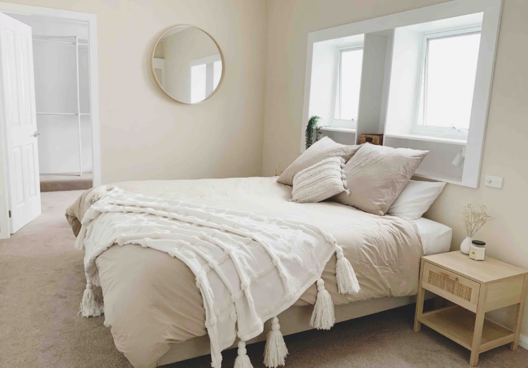How to Make a Boring Room Look Better: 15 Simple Fixes Neutral minimalist bedroom featuring soft beige bedding, a textured white tassel throw blanket, a round wall mirror, and a small wooden nightstand, showcasing warm design ideas on how to make a boring room look better.