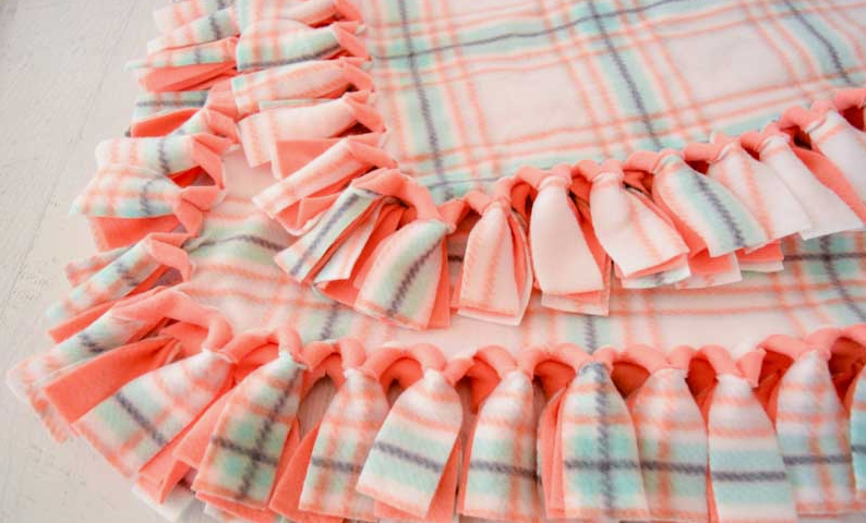 20 No-Sew DIY Crafts for Adults Close-up of a cozy fleece tie blanket featuring a peach and teal plaid pattern with hand-knotted fringe edges, demonstrating simple no-sew DIY crafts for adults.