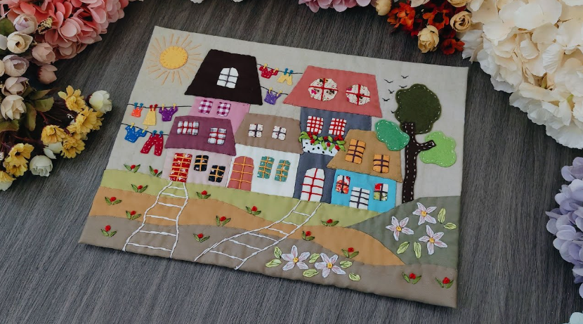 20 No-Sew DIY Crafts for Adults Fabric appliqué wall hanging depicting a whimsical village scene with colorful houses, trees, flowers, and a sun, showcasing creative no-sew DIY crafts for adults.