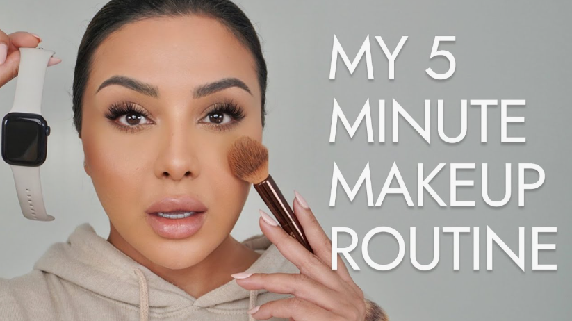 A woman holding up a smartwatch while applying powder to her cheek with a small makeup brush, demonstrating a quick 5 minute makeup routine.