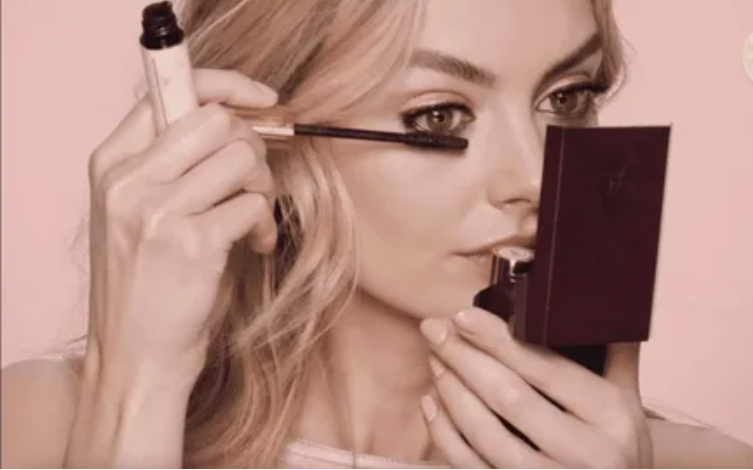 A woman holding a compact mirror while applying mascara to her eyelashes, highlighting a step in a quick 5-minute beauty routine.