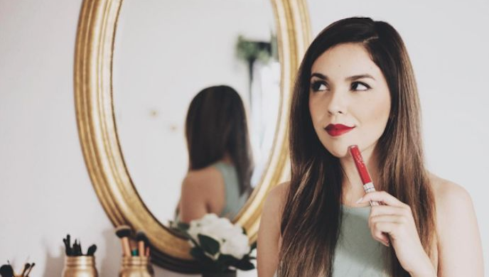 A woman holding a tube of red lipstick near her chin in front of a round gold mirror, completing a quick 5-minute beauty routine.