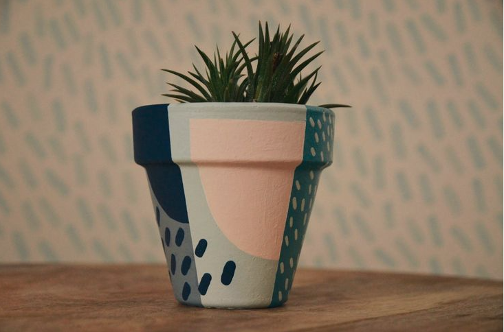 A painted terracotta flowerpot featuring an abstract geometric and dash pattern in shades of dark blue, peach, grey, and teal, holding a small green succulent.