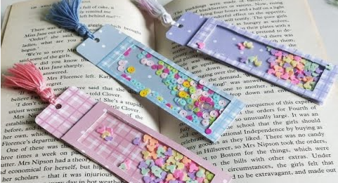 one hour crafts alt txt with keyphrase: Three handmade shaker bookmarks filled with colorful sequins and tiny decorative pieces resting on an open book, showcasing a creative project idea.