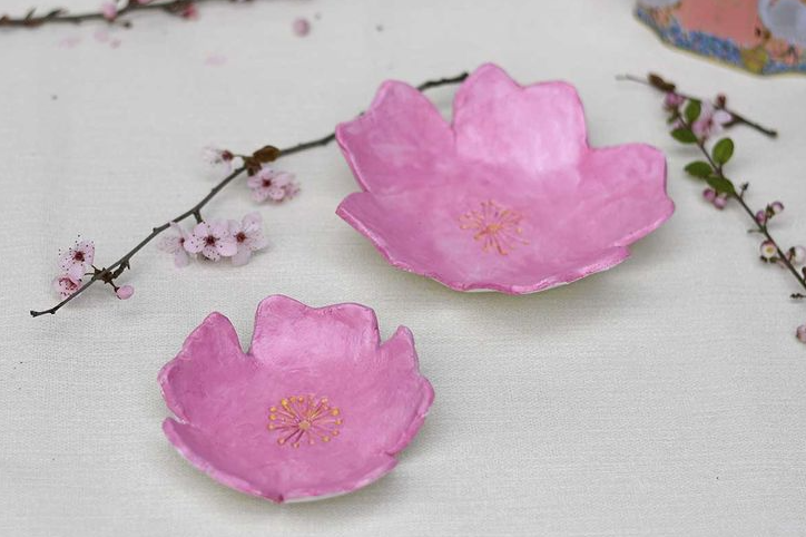 Two handcrafted pink ceramic flower dishes shaped like cherry blossoms, arranged on a light surface alongside delicate flowering branches, showcasing a creative project idea under the keyphrase one hour crafts.