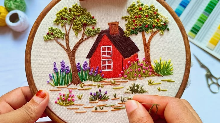 A hand embroidery project in a wooden hoop featuring a cozy red cottage surrounded by textured trees, flowers, and greenery, presented as a DIY craft under the keyphrase one hour crafts.