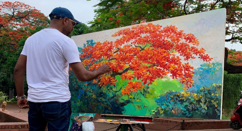 A male artist wearing a white t-shirt and a blue cap painting a large canvas outdoors, depicting a vibrant tree with bright orange-red blossoms against a green and blue background, created as part of an art project under the keyphrase one hour crafts.