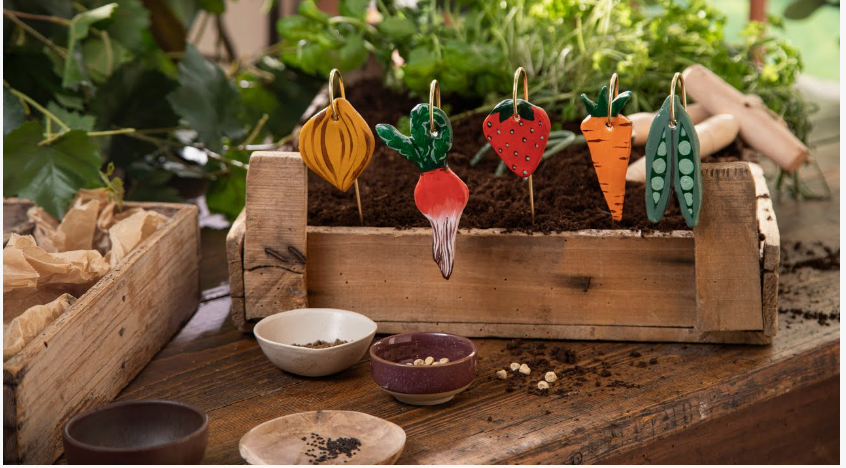 A set of handmade clay garden markers shaped and painted like vegetables including an onion, radish, strawberry, carrot, and pea pod, stuck into soil within a wooden planter box, showcased as a DIY craft under the keyphrase one hour crafts.