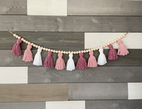 A decorative garland strung with natural wooden beads and alternating yarn tassels in shades of pink, dusty rose, mauve, and white, hung against a paneled wood wall as a DIY home decor project under the keyphrase one hour crafts.