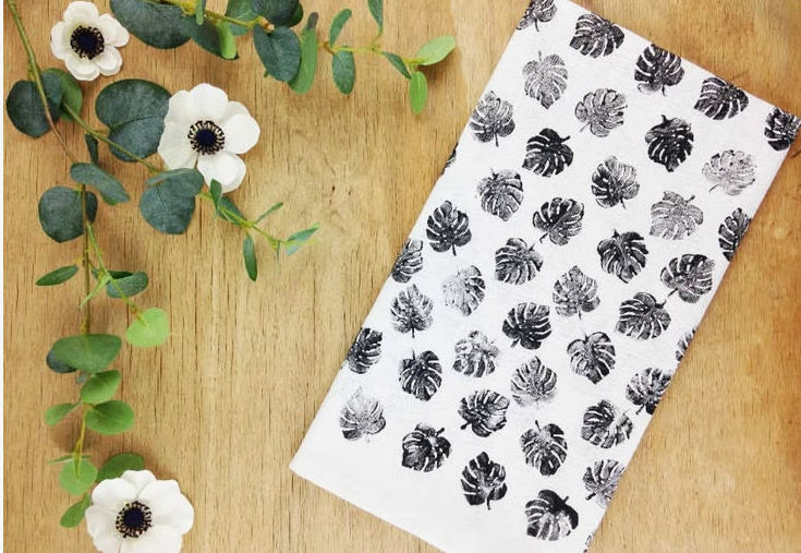 A hand-stamped white fabric tea towel featuring an all-over black monstera leaf pattern, displayed on a wooden surface next to faux white flowers and eucalyptus branches as a quick craft project under the keyphrase one hour crafts.