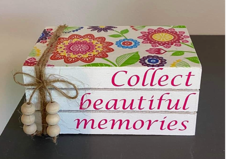 A stack of three decorative white wooden books stamped with the inspirational quote "Collect beautiful memories" in pink text, tied with twine and wooden beads, and adorned with a colorful floral pattern on top, presented as a DIY craft project under the keyphrase one hour crafts.