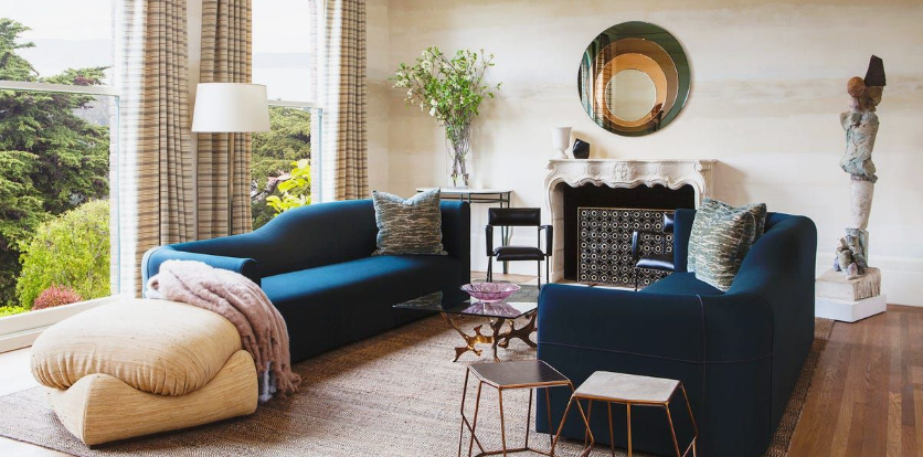 A stylish living room featuring two deep blue velvet sofas, a round statement mirror above a fireplace, a textured beige ottoman, and large windows looking out onto greenery, presented as an interior design inspiration project under the keyphrase one hour crafts.
