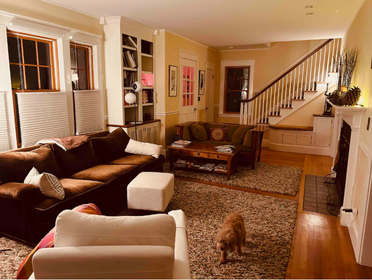 A cozy, warmly lit living room featuring a plush brown sofa, a cream-colored armchair, a wooden coffee table, built-in bookshelves, a staircase with a built-in bench, and a small dog walking across the rug, presented as a home styling project under the keyphrase one hour crafts.