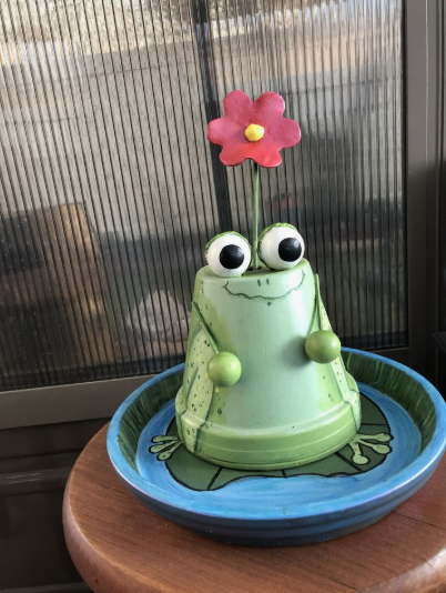 A whimsical green frog figurine holding a red flower, showcasing creative air dry clay ideas and painted pot crafts sitting on a blue lily pad tray.
