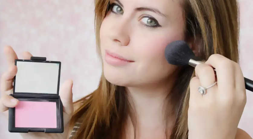 Beauty Products You Actually Need: A Minimalist Guide to Your Beauty Collection A woman applying pink blush with a large makeup brush while holding an open compact mirror showcasing a bright pink powder blush.