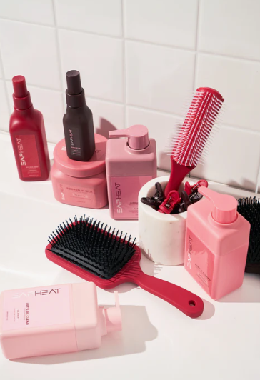 Beauty Products You Actually Need: A Minimalist Guide to Your Beauty Collection An arrangement of hair care essentials for beauty products you actually need, featuring pink and red bottles of shampoo, conditioner, styling products, hairbrushes, and sectioning clips against a tiled wall.