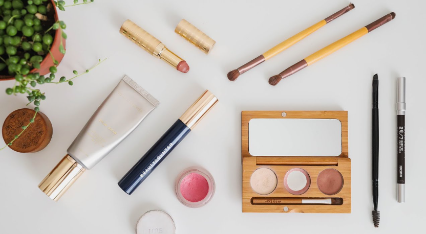 Beauty Products You Actually Need: A Minimalist Guide to Your Beauty Collection Flat lay arrangement of beauty products you actually need, featuring makeup brushes, a wooden eyeshadow palette, lipstick tubes, a tube of foundation, and eyeliner pencils on a clean white background next to a potted plant.