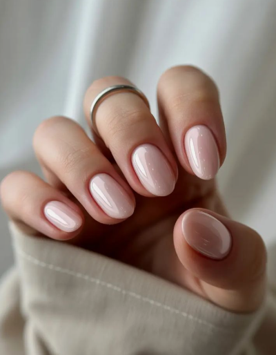 A close-up of a hand wearing a simple silver ring, showcasing elegant short nails for women over 50 with a glossy, milky nude gel manicure and neat, rounded shapes.