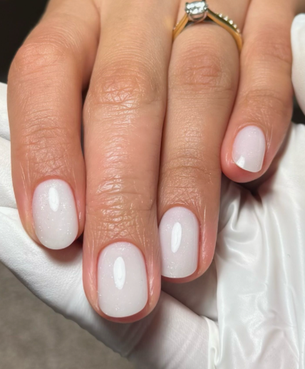 A close-up of a hand wearing a gold ring and a white glove, showcasing elegant short nails for women over 50 polished in a clean, milky white gloss.