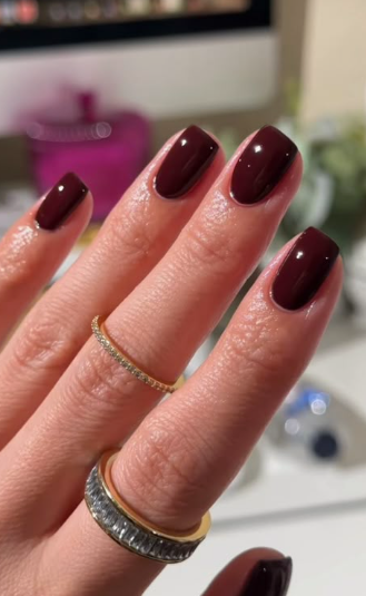 A close-up of a hand wearing two gold rings adorned with sparkling stones, showcasing elegant short nails for women over 50 polished in a glossy, dark oxblood red.