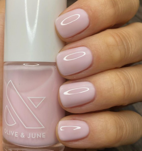 A close-up of a hand holding an Olive & June polish bottle, showcasing elegant short nails for women over 50 polished in a glossy, milky light pink.